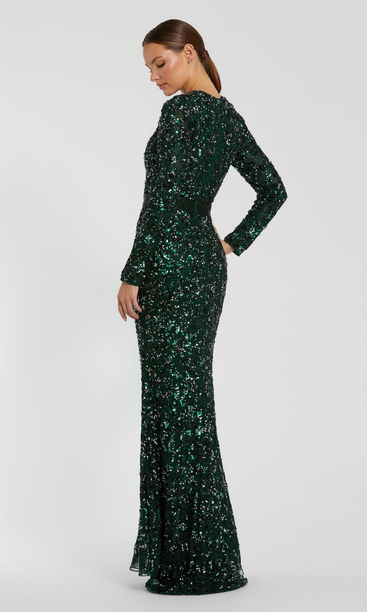 A woman stands in profile against a plain light background, shimmering in the Mac Duggal 5453 Long Sleeve Sequin Formal Dress—floor-length, dark green, and covered in sparkling sequins.